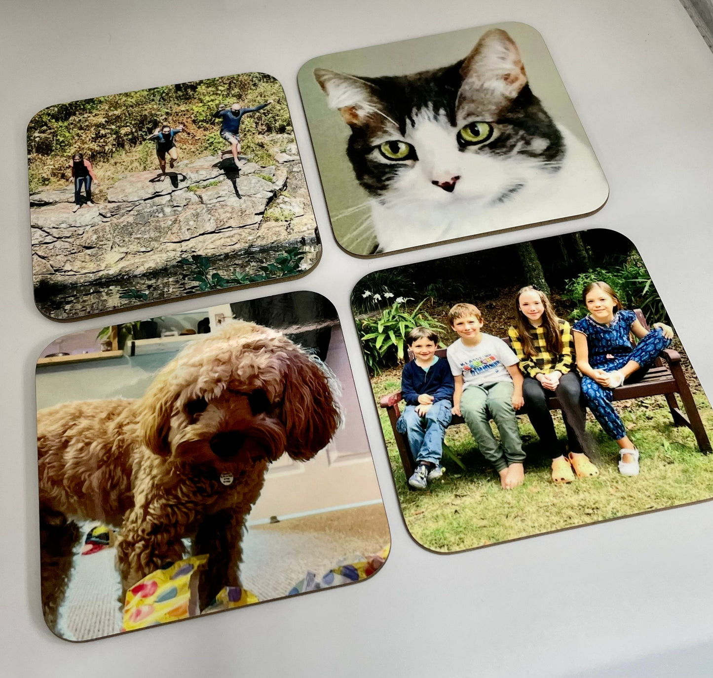 Custom coasters showing 4 different photo designs of a cat, a dog, a family photo and an adventure photo of 3 people jumping in to a river